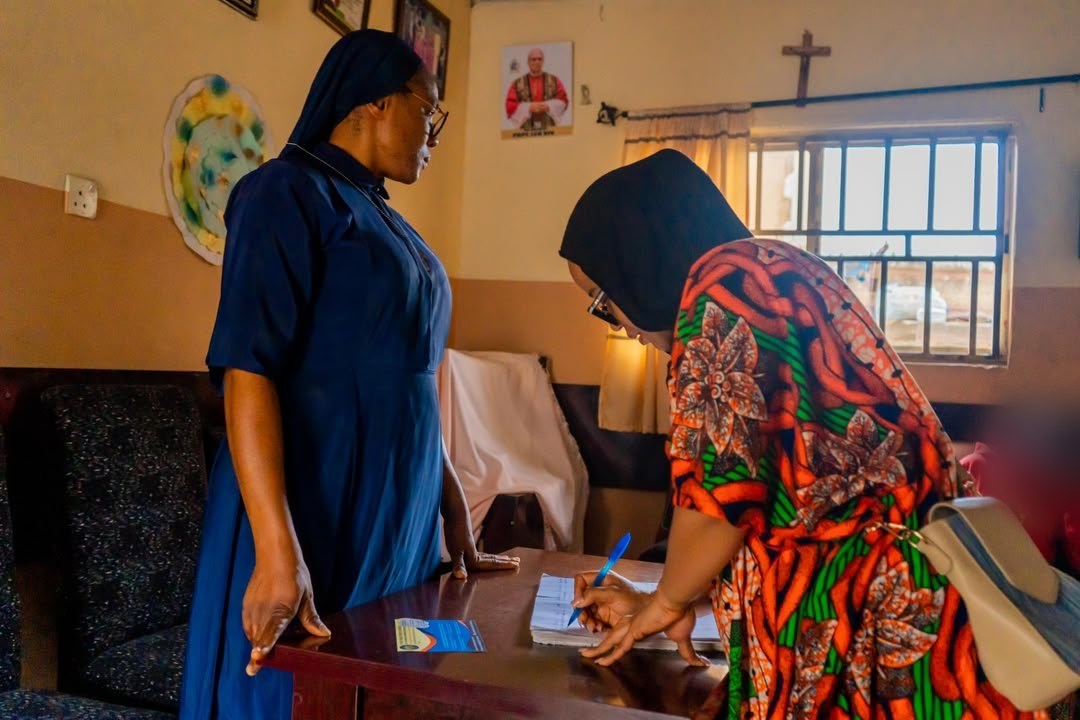 Anita Ejumudo writing at a desk inside the Home as one of the Sisters looks on